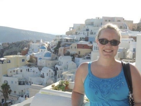 White woman with sunglasses standing in front of a sun drenched, cliff side town. 